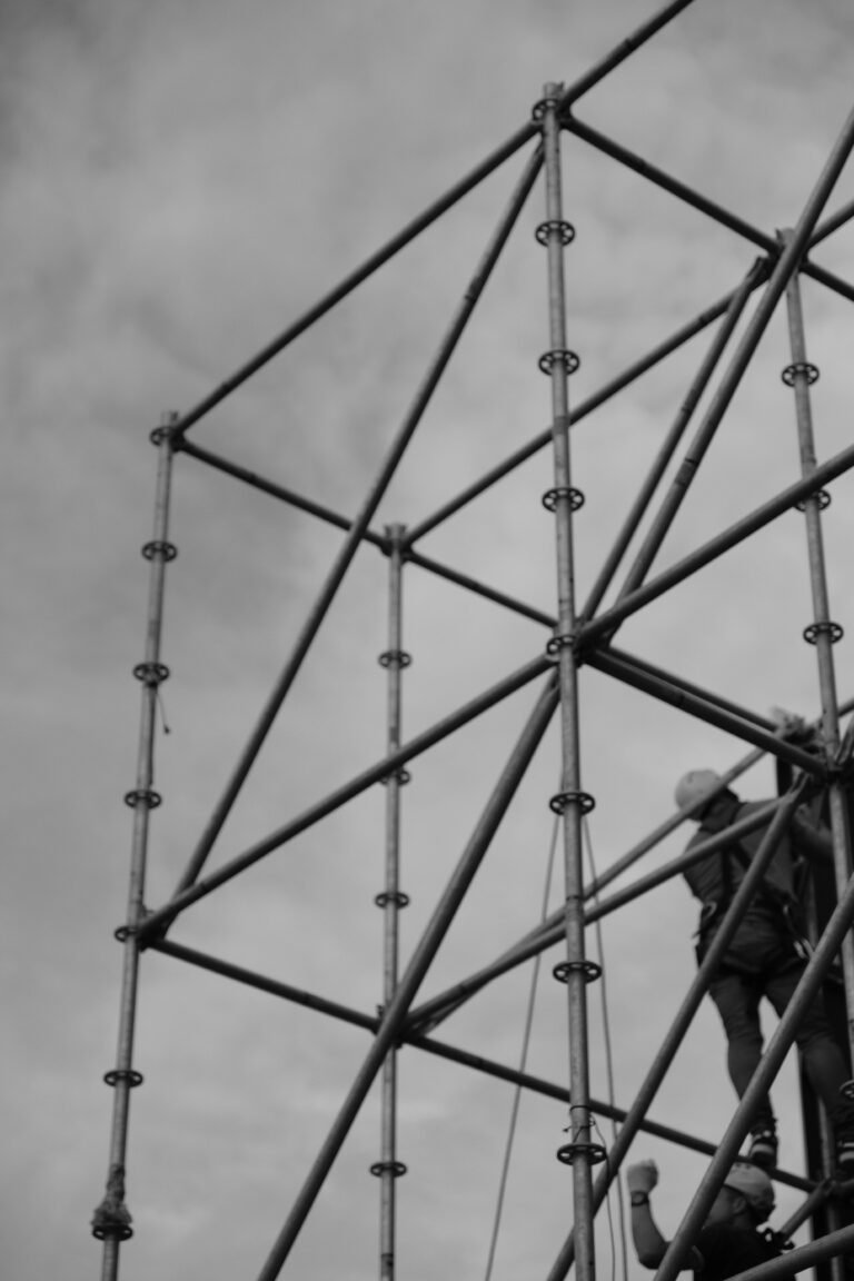 Black and white image of a worker on scaffolding set against a cloudy sky.